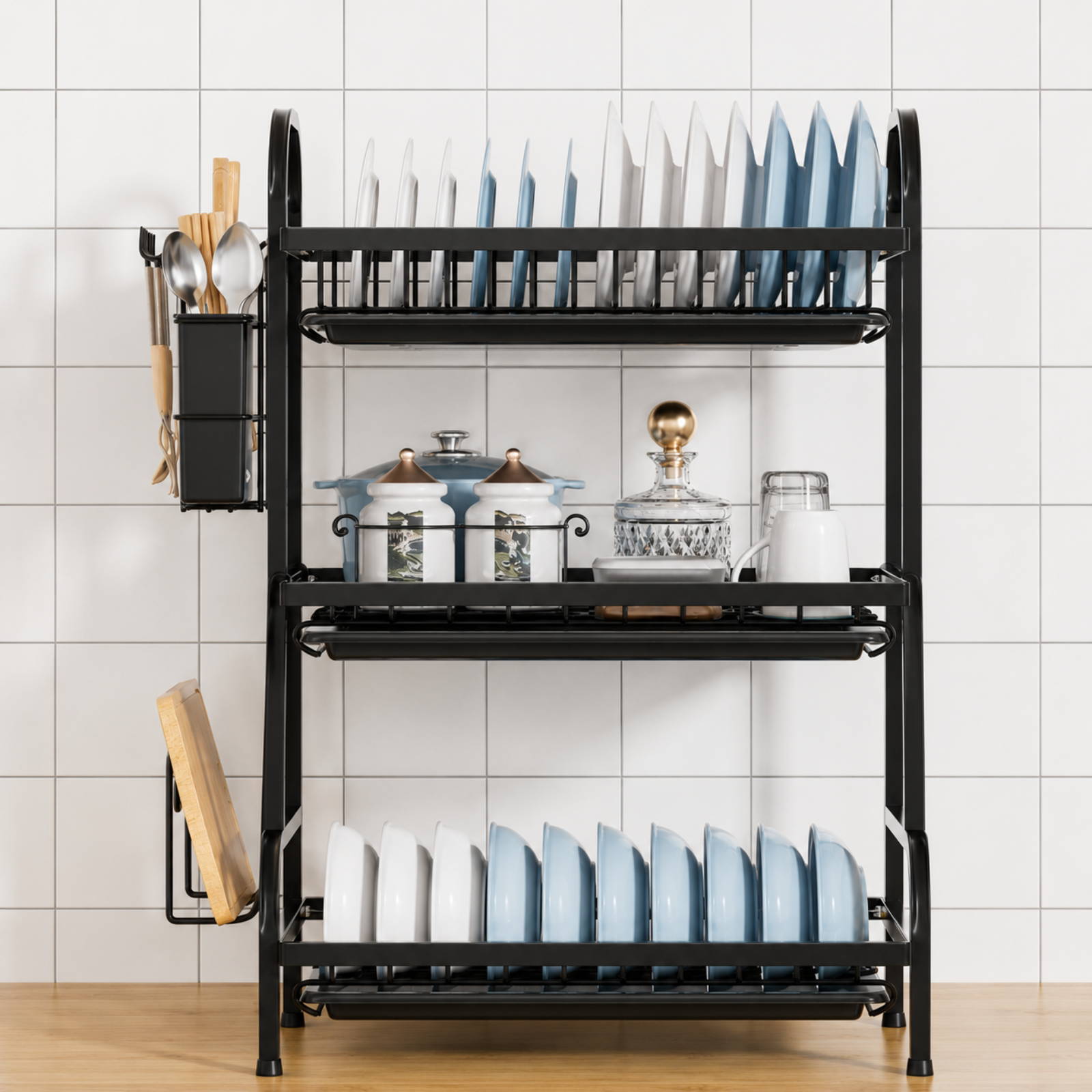 A black metal 3-tier kitchen dish rack with a side hanging utensil holder, shown stocked with plates, jars, and cookware against a white tiled wall.