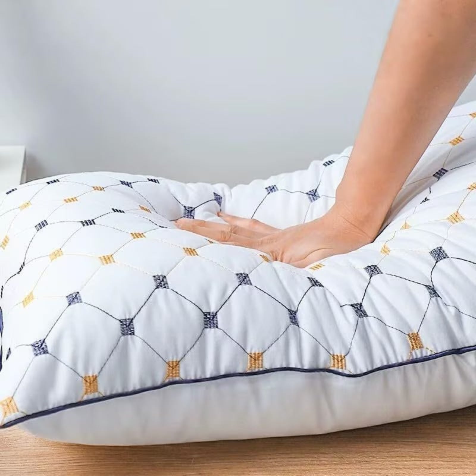 A white quilted lumbar pillow with a grey and yellow geometric lattice pattern, shown with a person's hand pressing down on the canter to demonstrate its soft, supportive cushioning.