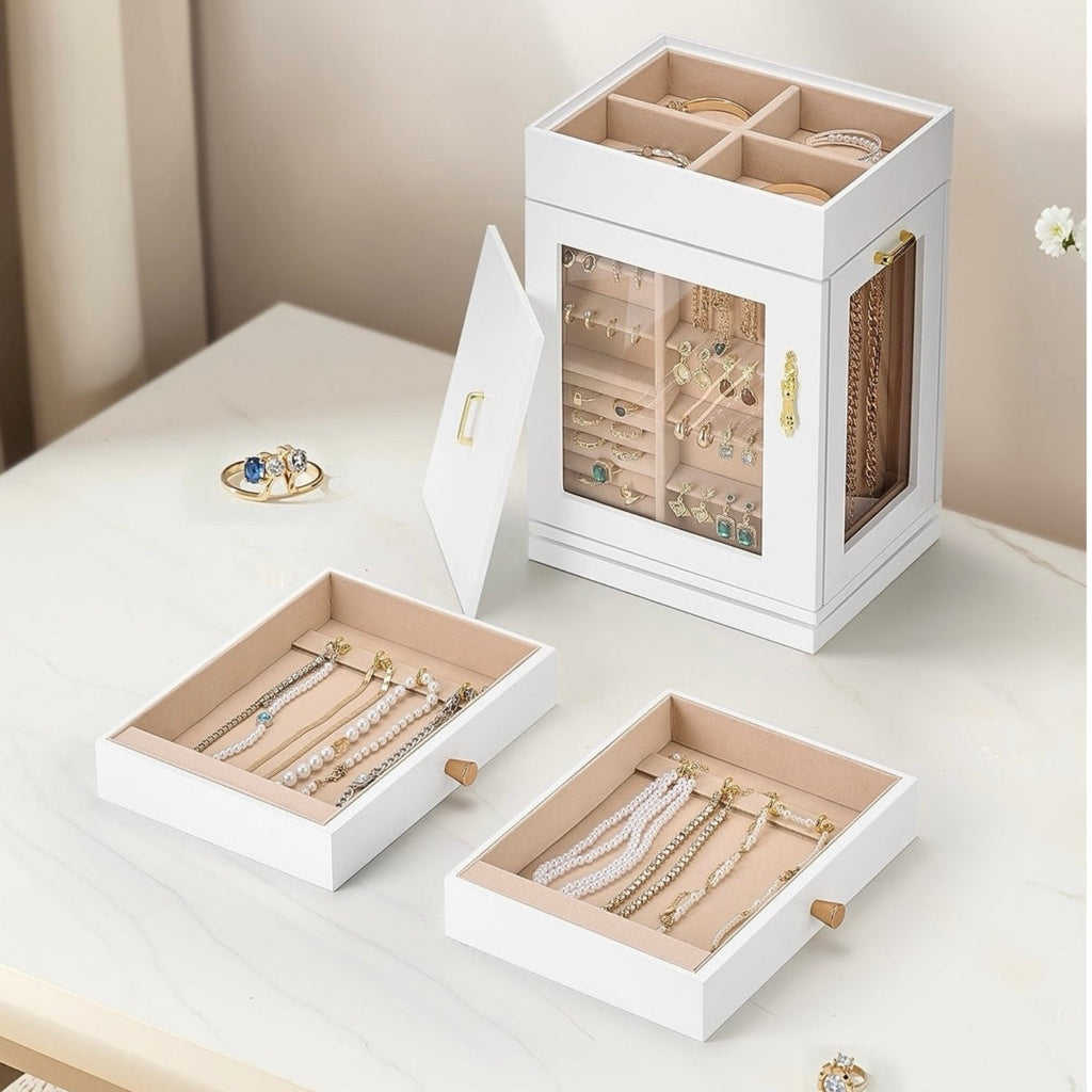 A white jewelry organizer box shown open, with two drawers pulled out below and a glass-front upper compartment displaying jewelry. The scene is styled on a light tabletop with a soft beige background and no people or animals present.