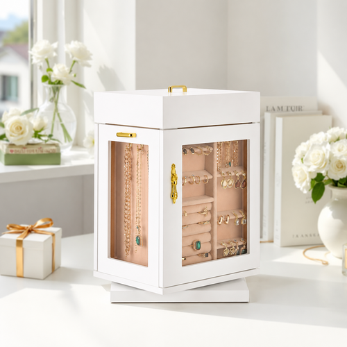 A white jewelry organizer box with glass doors and gold-tone handles, shown on a tabletop in a bright, styled room. The organizer contains multiple layers of necklaces, bracelets, and earrings in soft blush and gold tones.