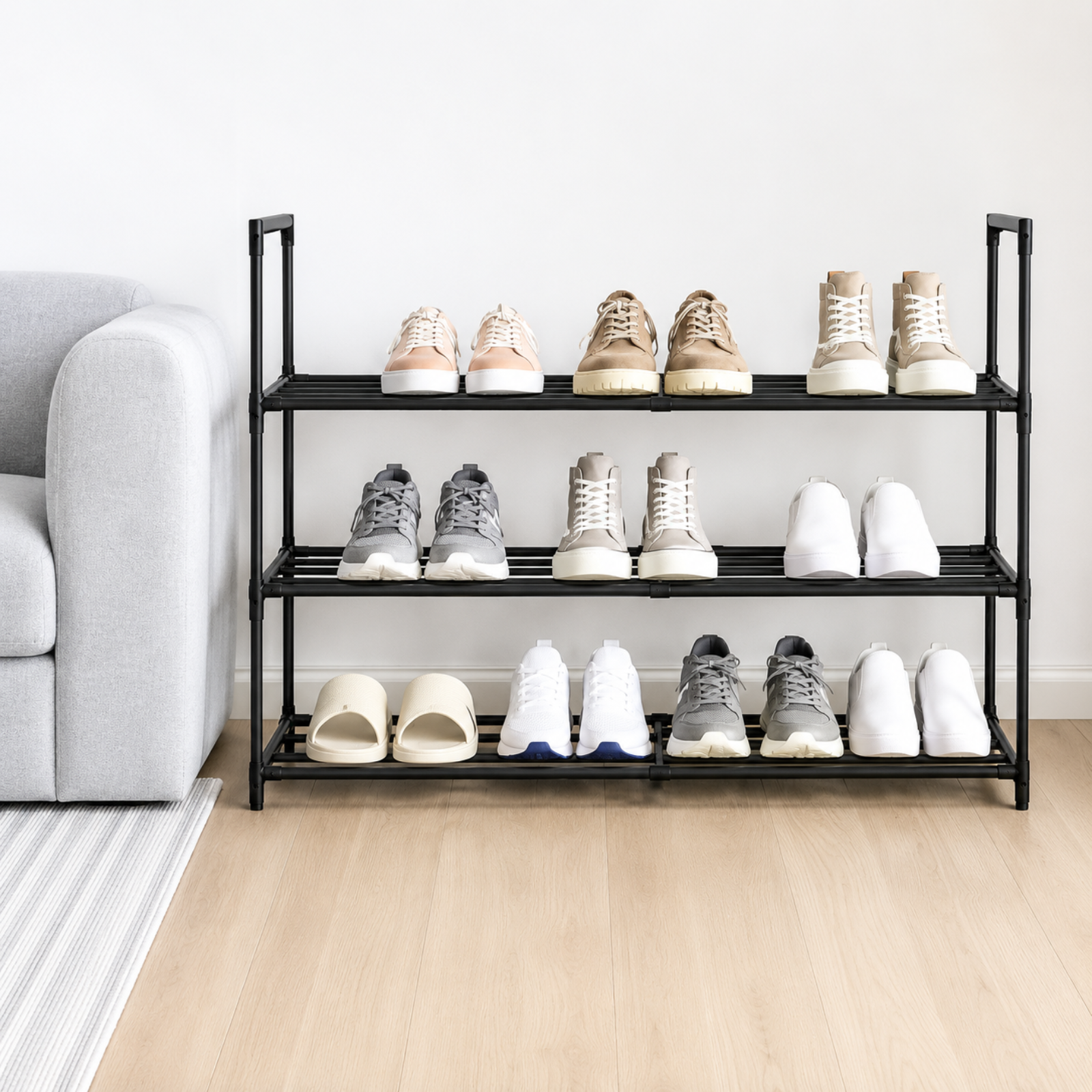 A three-tier black metal shoe rack holding assorted sneakers, boots, and slippers, staged in a bright living room next to a light gray sofa on a light wood floor.