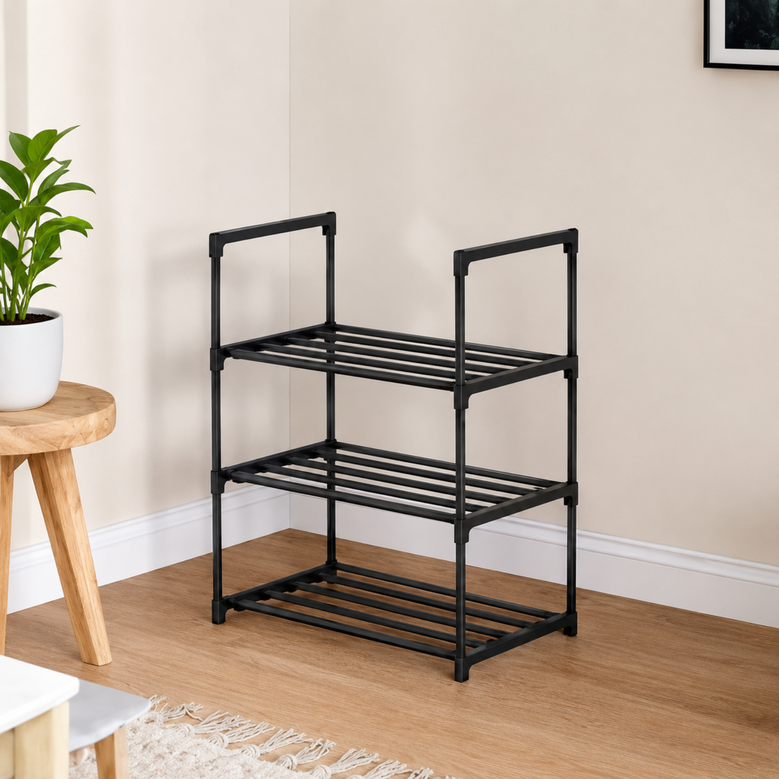 A three-tier black metal shoe rack with integrated side handles, shown in a home entryway setting with a wooden floor, beige wall, and a small wooden stool with a potted plant.