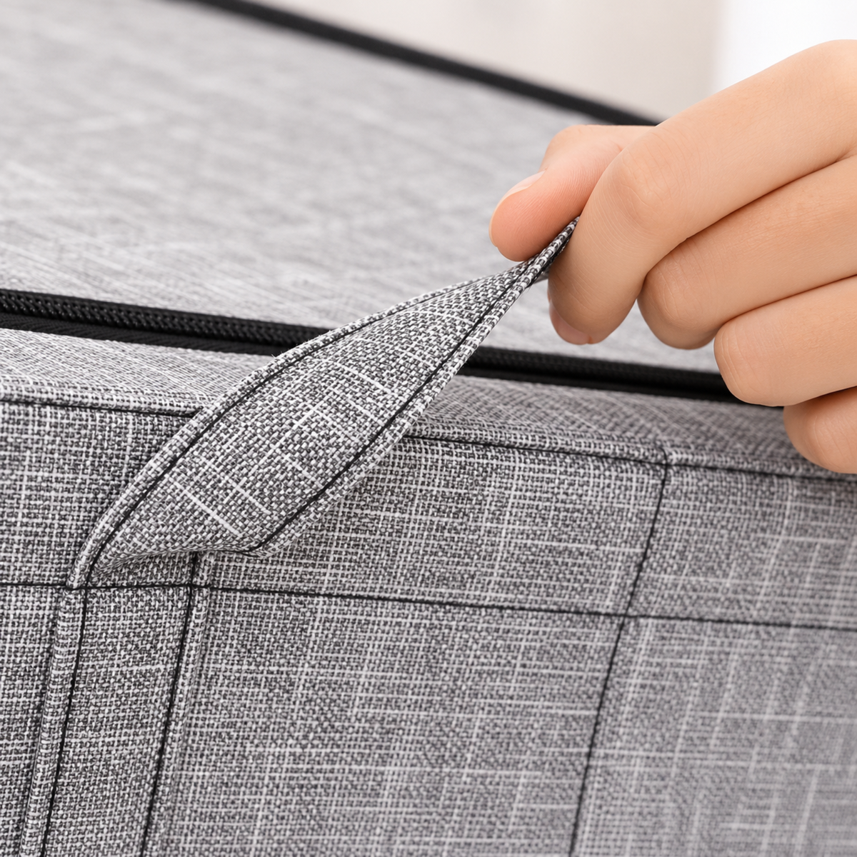 Close-up of a hand lifting a corner of a gray textured fabric storage bin to show the zipper and material detail.