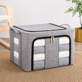 A gray fabric storage bin with a zippered front pocket and side handles, shown on a woven rug in a styled room setting with a chair and plant.