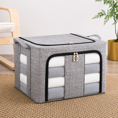 A gray fabric storage bin with a zippered front pocket and side handles, shown on a woven rug in a styled room setting with a chair and plant.