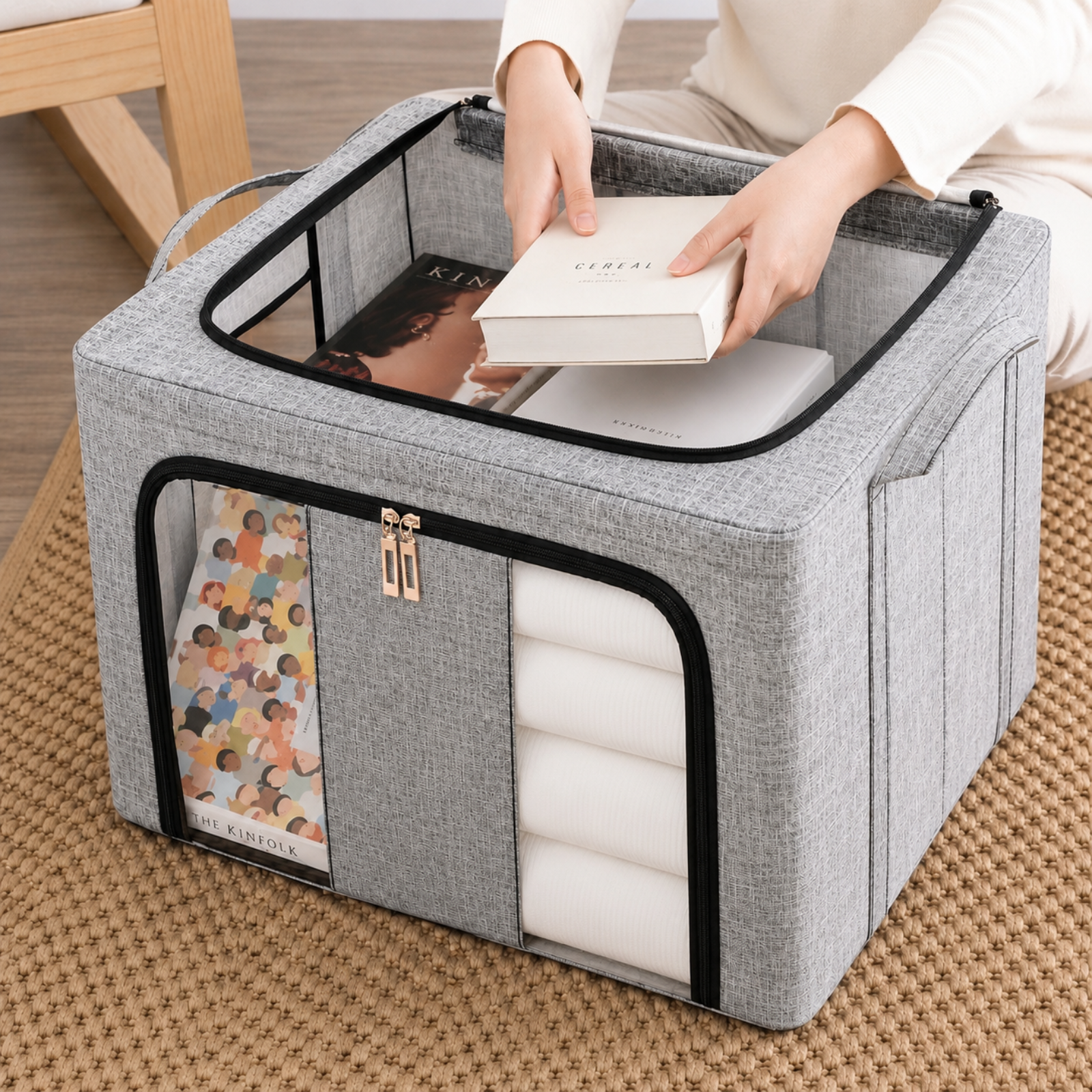 A gray fabric storage bin with clear zippered windows, shown on a woven rug with a person's hands placing a white box inside.