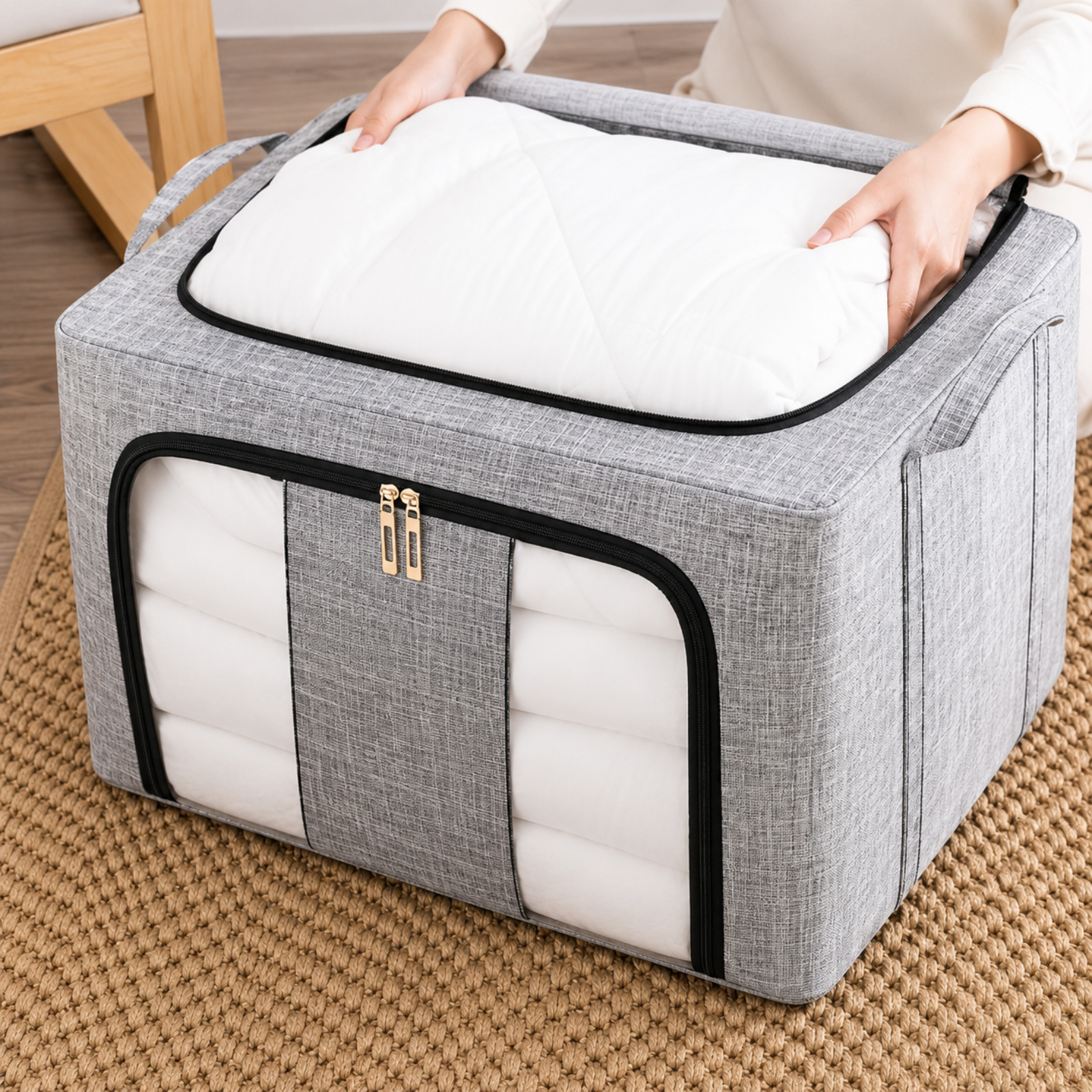 A gray fabric storage bin with black trim and gold zipper pulls, shown with a person's hands opening the top lid to reveal white quilted bedding or cushion inserts inside, placed on a woven tan rug.