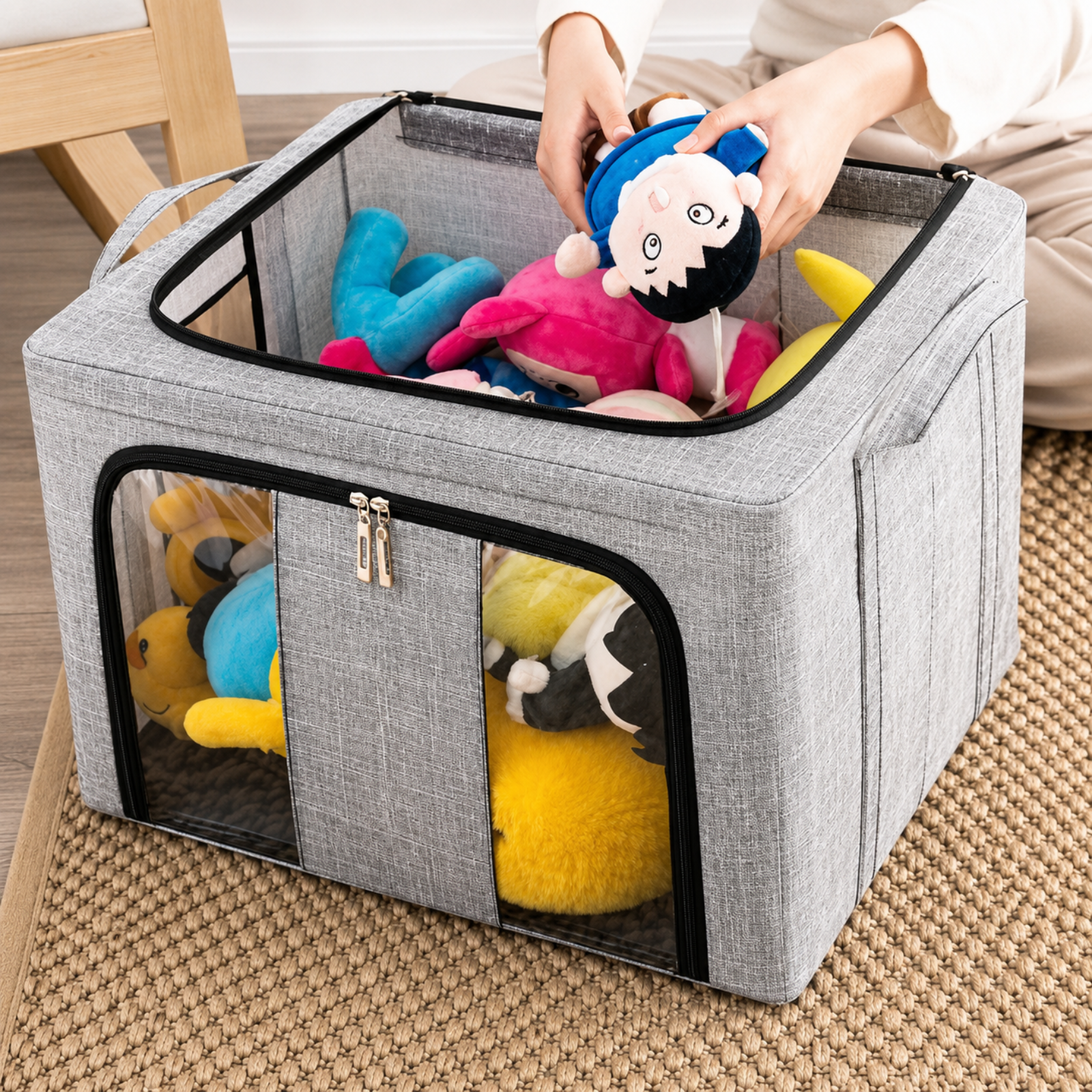 A gray fabric storage bin with clear front window panels, shown on a woven rug with stuffed toys inside. A person's hands are placing a plush doll into the top compartment, demonstrating use in a home setting.