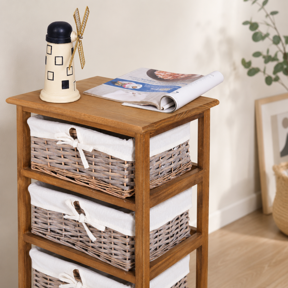 A wooden three-tier side table with woven wicker baskets lined with white fabric on each shelf, styled with a small decorative windmill bottle and an open magazine on top, in a softly lit room.