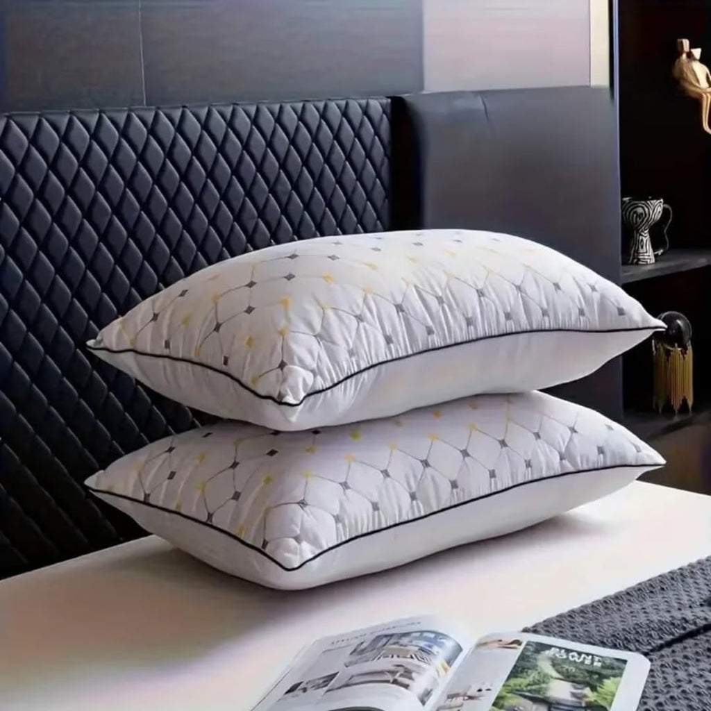 Two white quilted pillows stacked on a white table, featuring a geometric diamond pattern in grey and yellow with black piping, set against a dark quilted headboard.