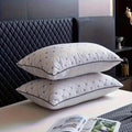 Two white quilted pillows stacked on a white table, featuring a geometric diamond pattern in grey and yellow with black piping, set against a dark quilted headboard.