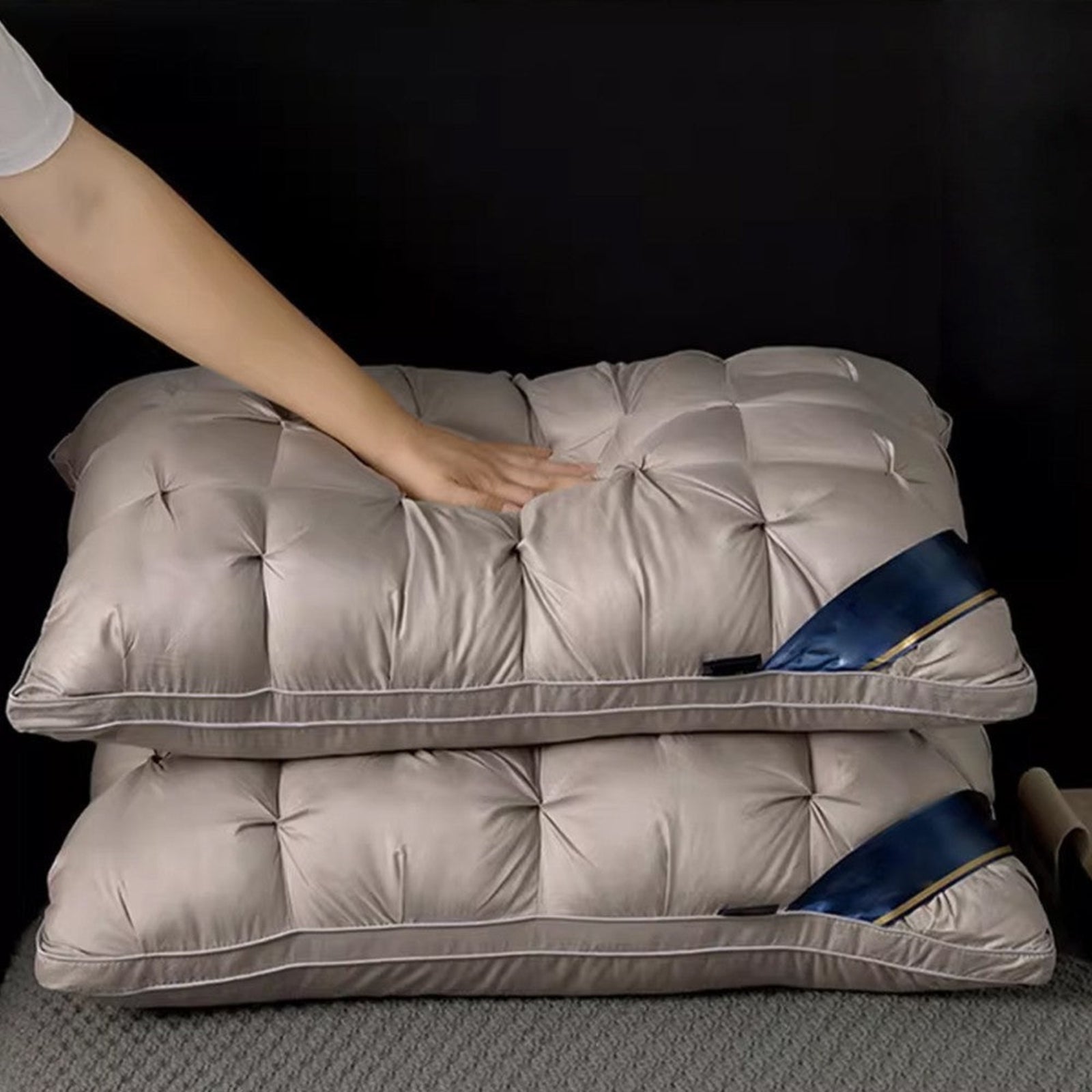 Two padded quilted pillows in a light taupe-beige color stacked on a textured gray surface against a black background, with a person's hand pressing down on the top pillow and a blue fabric tag visible on the side.