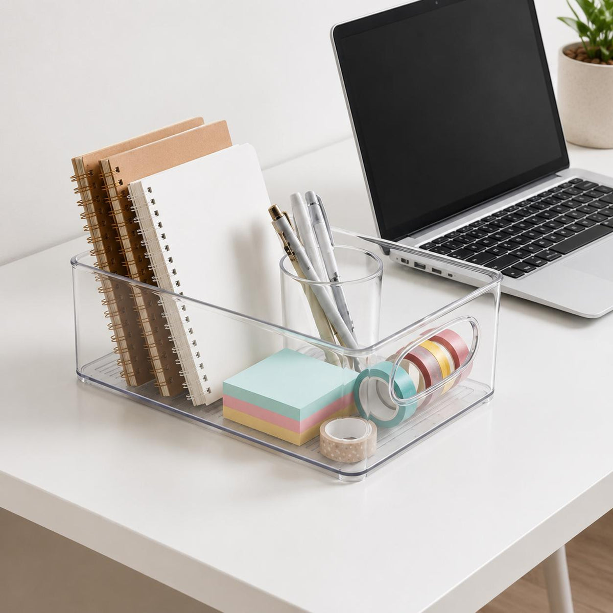 Clear plastic desk organizer tray on a white desk, holding notebooks, pens in a glass cup, sticky notes, and washi tape, with a laptop and plant in the background.