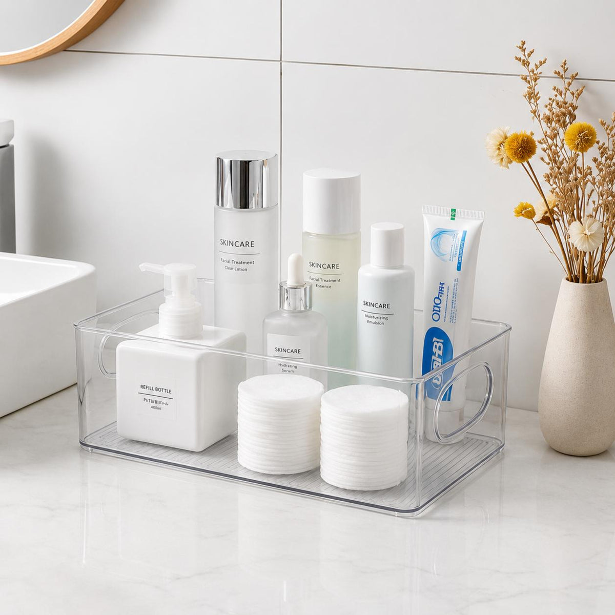 A clear plastic bathroom organizer tray holding several skincare and beauty products, including a pump bottle, tubes, and cotton pads, set on a white marble bathroom counter.