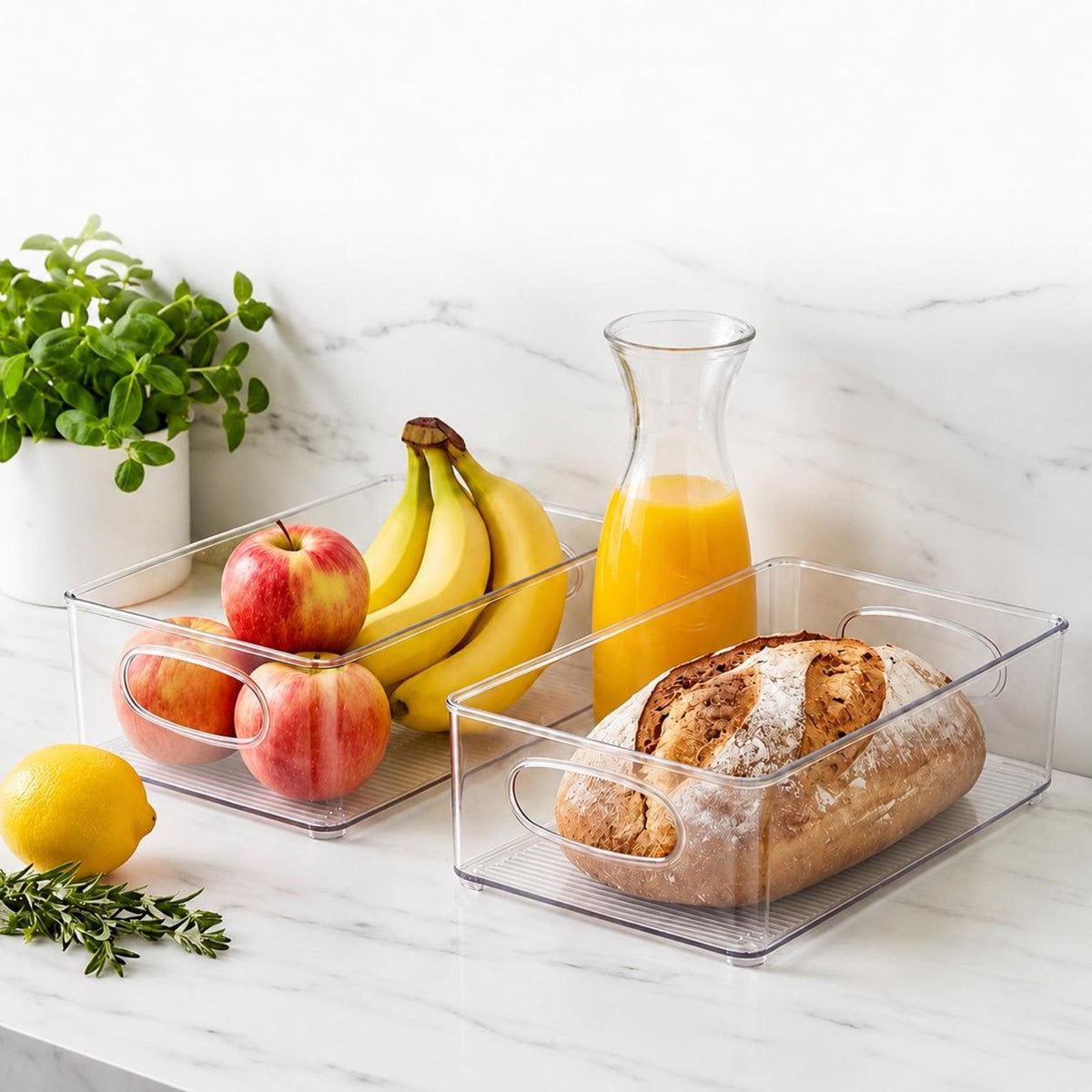 Two clear plastic food storage bins on a white marble countertop, filled with fruit, a loaf of bread, and a glass carafe of orange juice, with a potted plant in the background.
