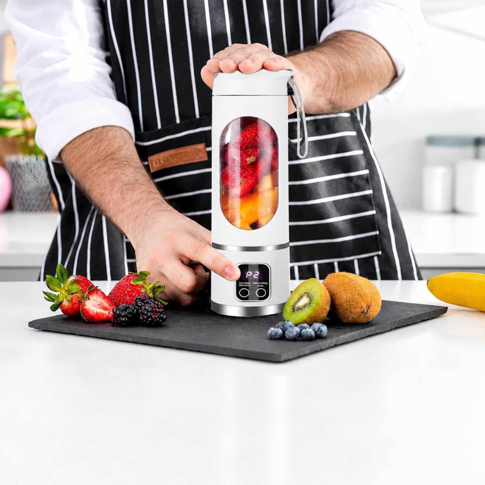 A person in a white shirt and black-and-white striped apron holds a portable electric blender with a clear fruit jar on a black cutting board, surrounded by strawberries, kiwi, blueberries, and a banana in a bright kitchen setting.