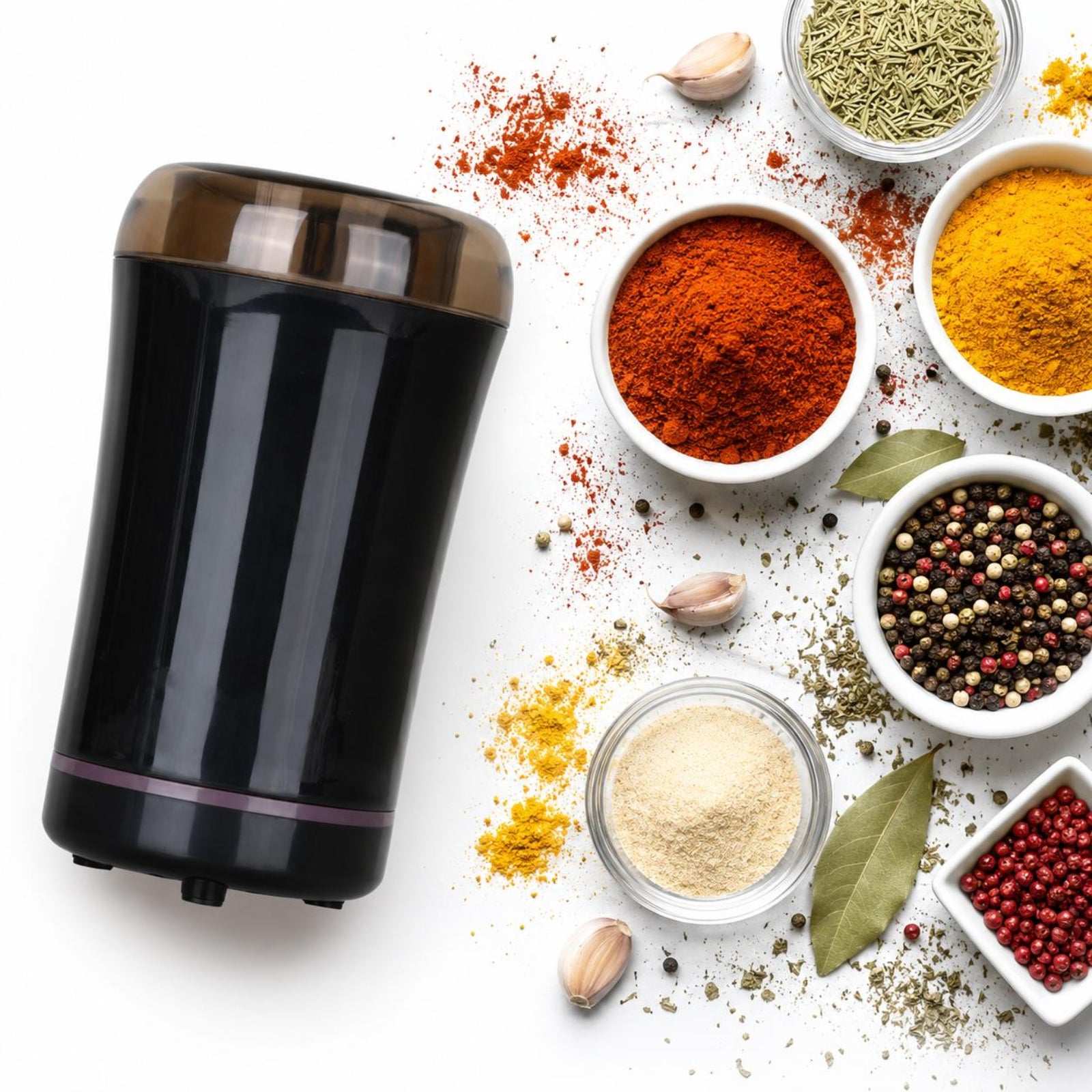 A black electric spice grinder with a clear lid is shown on a white background, surrounded by various whole and ground spices in small bowls, including paprika, turmeric, cumin, garlic, bay leaves, and peppercorns.