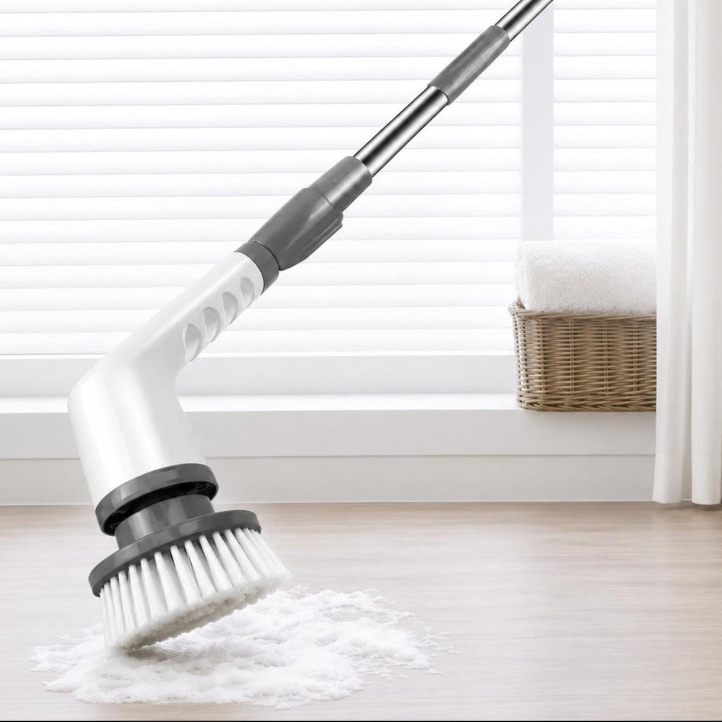 A flexible floor scrubber with a white and gray handle and bristle head, shown on a light wood floor with a pile of cleaning powder in front of a window with blinds and a curtain.