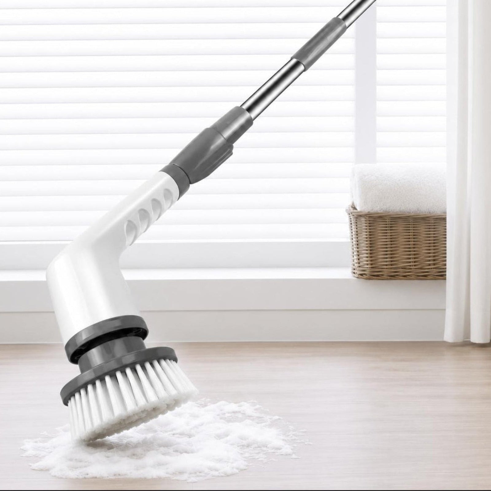 A flexible floor scrubber with a white and gray handle and bristle head, shown on a light wood floor with a pile of cleaning powder in front of a window with blinds and a curtain.